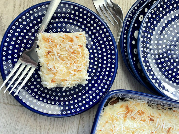 A slice of coconut cake on a small dark blue white polka-dotted plate with a fork lying on the plate. In the bottom right corner is a corner of a dark blue 9x13 baking dish with three small dark blue polka-dotted stacked plates above it.
