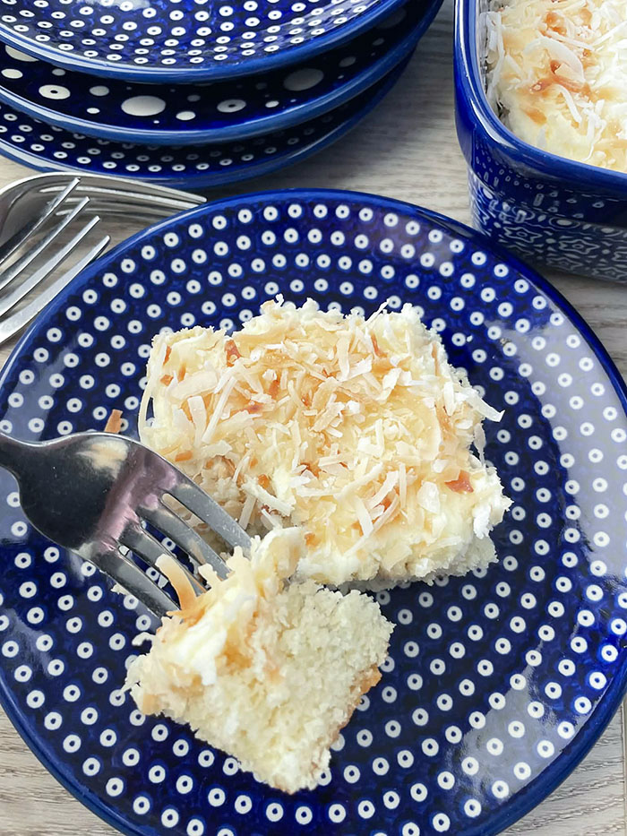 A slice of coconut cake with a corner of the slice cut out with a for inserted into it from the left on a small dark blue plate with polka dots. In the upper left corner are two fork heads and partial picture of three small stacked plates. In the upper right corner is the edge of a 9x13 baking dish.