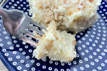 A small dark blue white polka-dotted Polish pottery plate with a slice of coconut cake on it with a fork held from the left inserted into a corner piece of the slice of cake.