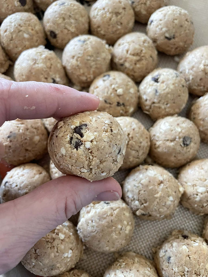 A protein power ball held between a pointer finger and a thumb above a baking sheet lined with a silpat mat filled with protein power balls.