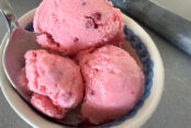 A bowl of raspberry ice cream with three scoops. A spoon is inserted diagonally into the bowl from the left.