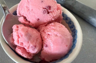 A bowl of raspberry ice cream with three scoops. A spoon is inserted diagonally into the bowl from the left.