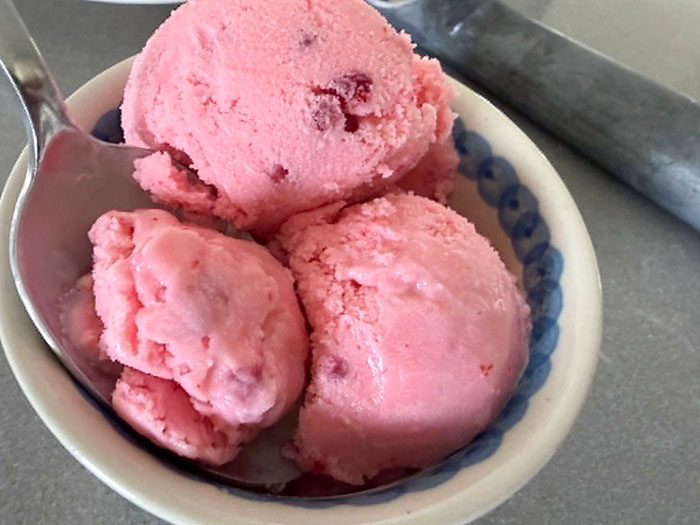 A bowl of raspberry ice cream with three scoops. A spoon is inserted diagonally into the bowl from the left.