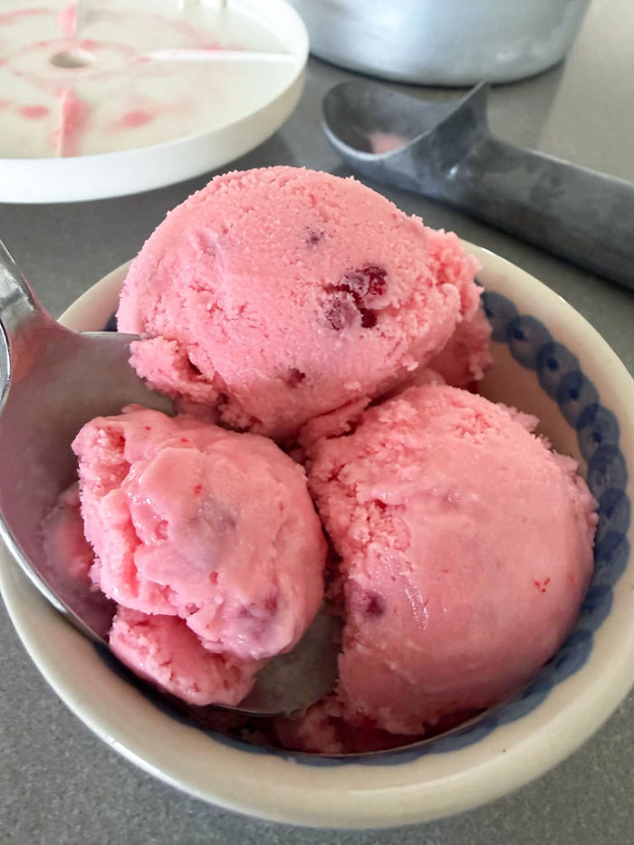 A bowl of three scoops of Homemade Raspberry Ice Cream with a spoon inserted diagonally from the left. Faded in the background is an ice cream scoop and a lid.