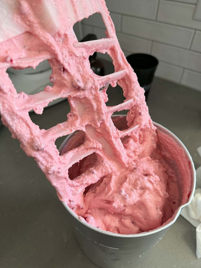 A canister of Raspberry Ice Cream with the ice cream paddle pulled almost all the way out with ice cream on it.