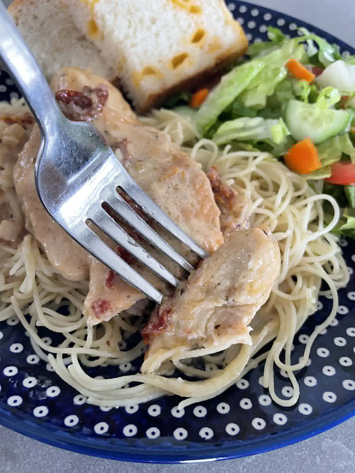 A dark blue plate with white polka dots with Marry Me Chicken on a bed of angel hair pasta. A green salad with cucumbers and carrots, and a couple of slices of cheesy bread are also on the plate.