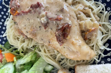 A piece of Marry Me Chicken on top of angel hair pasta on a dark blue and white polka dotted plate with a green salad, and slices of bread.