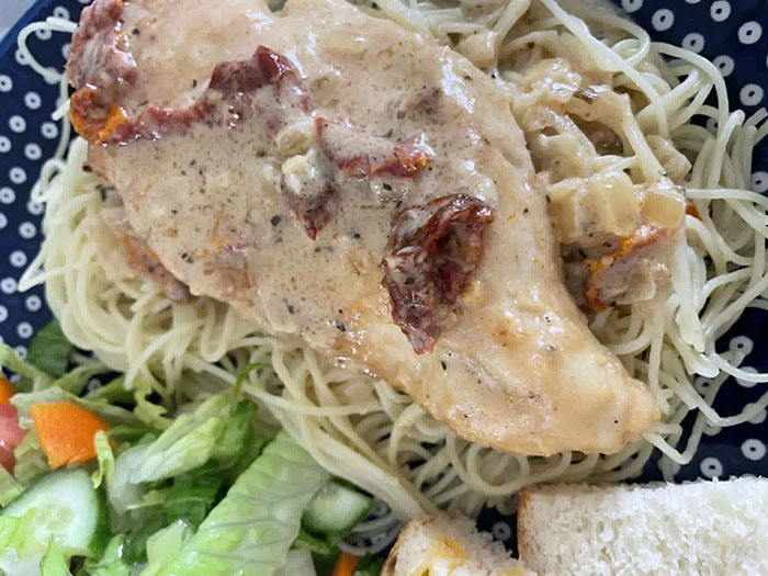 A piece of Marry Me Chicken on top of angel hair pasta on a dark blue and white polka dotted plate with a green salad, and slices of bread.