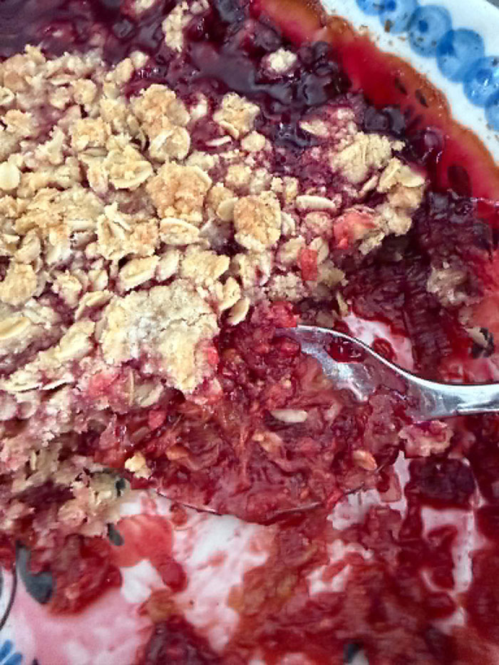 A baking dish of raspberry rhubarb crisp with a serving scooped out and a spoon with crisp on it lying in the dish from the right.