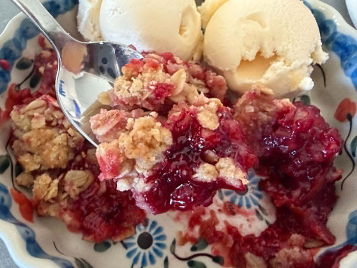 A serving of raspberry rhubarb crisp with two scoops of vanilla ice cream with a spoon held above diagonally from the left with some crisp on it.