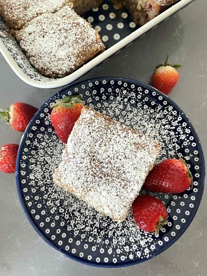 A square piece of strawberry rhubarb cake dusted with powdered sugar on a small dark blue white polka-dotted plate dusted with powdered sugar with two strawberries on the right of the piece of cake and one on the left. A few strawberries are surrounding the top of the plate. The corner of a 9x13 baking dish of a sliced strawberry rhubarb cake is in the upper left middle.