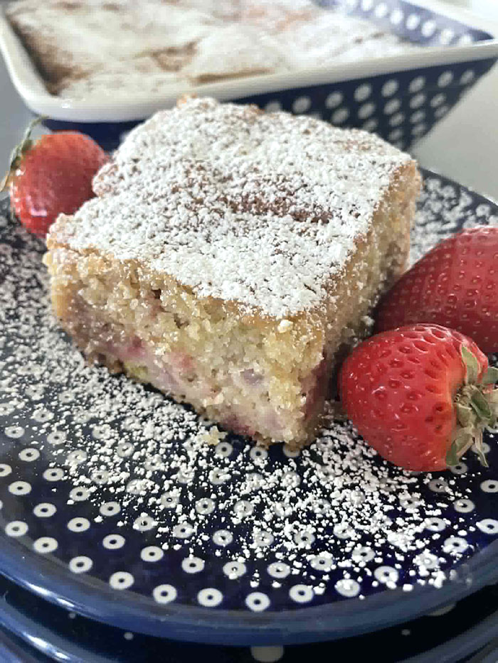 A small stack of dark blue, white polka-dotted small plates with the top plate having dusting of powdered sugar and a slice of strawberry rhubarb coffee cake with two strawberries on the right of the cake and one strawberry on the left of the cake. Faded in the background is a 9x13 baking dish of Strawberry Rhubarb Coffee Cake.
