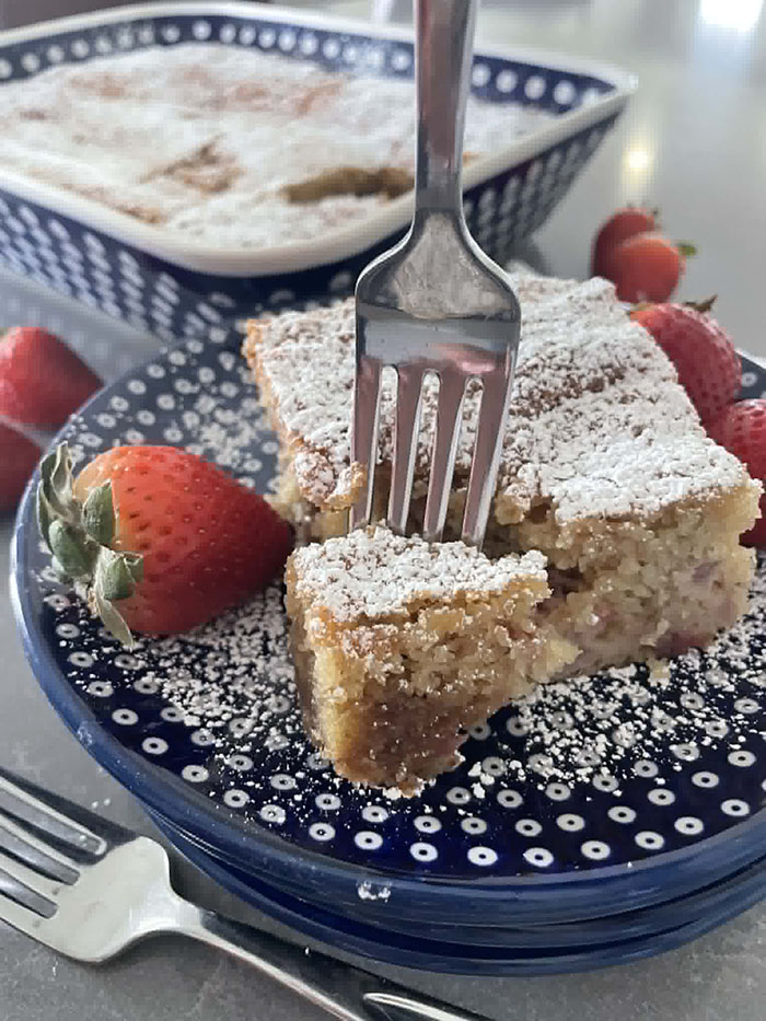 A slice of Strawberry Rhubarb Coffee Cake dusted with powdered sugar on a stack of dark blue and white polkadotted plates surrounded by whole strawberries with a fork inserted vertically into a corner of the slice of cake. In the lefthand bottom corner is a fork, and faded in the background is a 9x13 baking dish of Strawberry Rhubarb Coffee Cake.