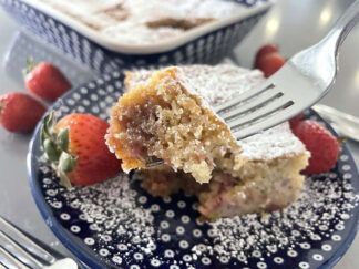 A square slice of strawberry rhubarb coffee cake sitting on a dark blue white polka-dotted dessert plate dusted with powdered sugar and a couple of whole strawberries. A corner of the cake is held up with a fork from the right. Faded in the background are whole strawberries around the corner of a 9x13 dish of Strawberry Rhubarb Coffee Cake.