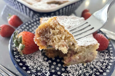 A square slice of strawberry rhubarb coffee cake sitting on a dark blue white polka-dotted dessert plate dusted with powdered sugar and a couple of whole strawberries. A corner of the cake is held up with a fork from the right. Faded in the background are whole strawberries around the corner of a 9x13 dish of Strawberry Rhubarb Coffee Cake.