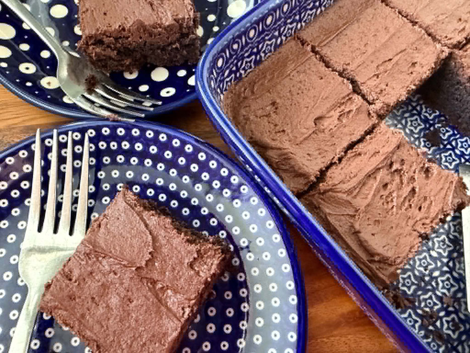 A frosted brownie on a dark blue plate with white polka dots with a fork lying next to the brownie on the plate. To the right of the plate is the corner of a 9x13 baking dish of sliced frosted brownies. In the upper left corner is a partial picture of another brownie on a dark blue plate with polka dots and a fork on the plate next to the brownie.
