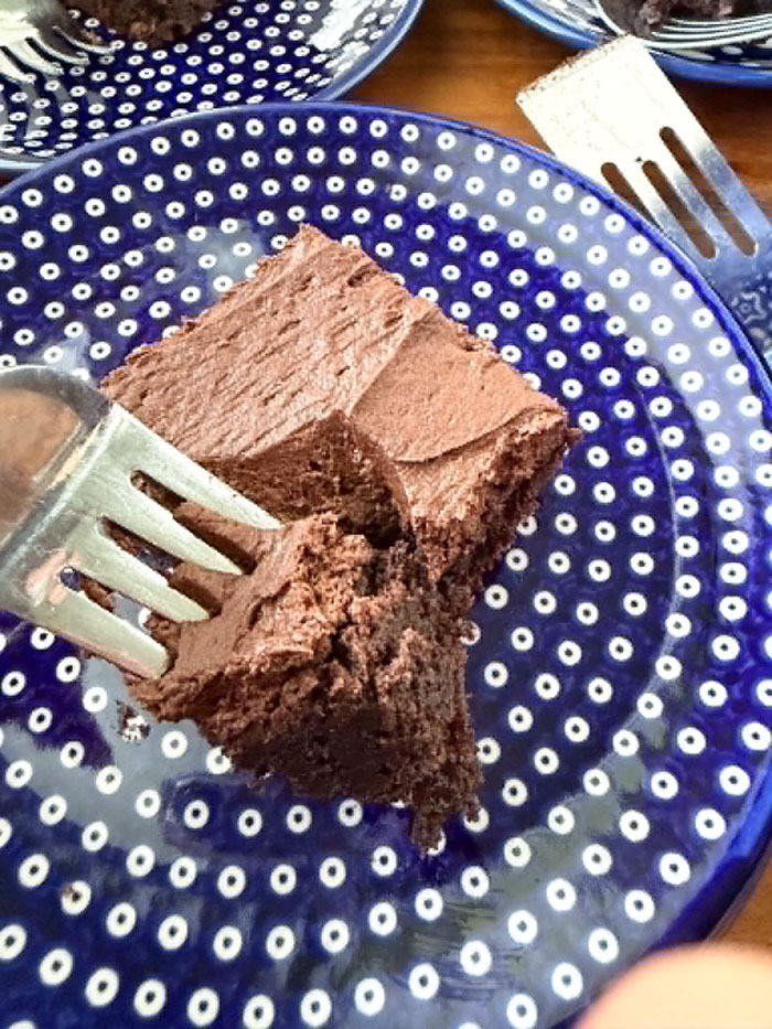 A small dark blue Polish pottery plate with white polka dots with a frosted brownie in the middle. A fork is held diagonally from the left of the plate with a bite on it pulling away from a corner of the frosted brownie. In the upper background are partial pictures of another small plate and the head of a serving spatula.