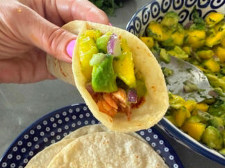 A blackened salmon taco with mango avocado salsa held above a plate of corn tortillas and a bowl of mango avocado salsa.