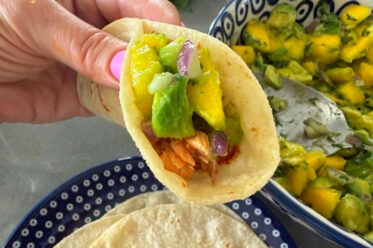 A blackened salmon taco with mango avocado salsa held above a plate of corn tortillas and a bowl of mango avocado salsa.