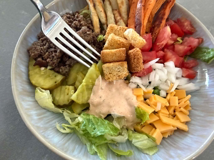 A bowl of chopped lettuce topped with burger sauce, small slices of cheddar cheese, diced onion, diced tomatoes, sweet potatoe fries, french fries, ground beef, croutons, and sliced pickle chips. A fork is lying diagonally, partially on the bowl in the upper left corner.
