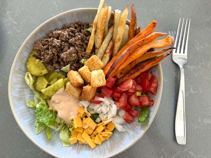 A bowl with chopped lettuce on the bottom topped with sliced pickle chips, ground beef, french fries, sweet potato fries, diced tomatoes, diced onion, small slices of cheese, smash burger sauce, and croutons. On the right side of the bowl is a fork.