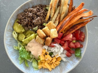 A bowl with chopped romaine lettuce topped with small slices of cheese, diced onions, diced tomatoes, sweet potato fries, french fries, ground beef, sliced pickle chips, burger sauce, and croutons.
