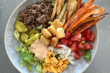 A bowl with chopped romaine lettuce topped with small slices of cheese, diced onions, diced tomatoes, sweet potato fries, french fries, ground beef, sliced pickle chips, burger sauce, and croutons.