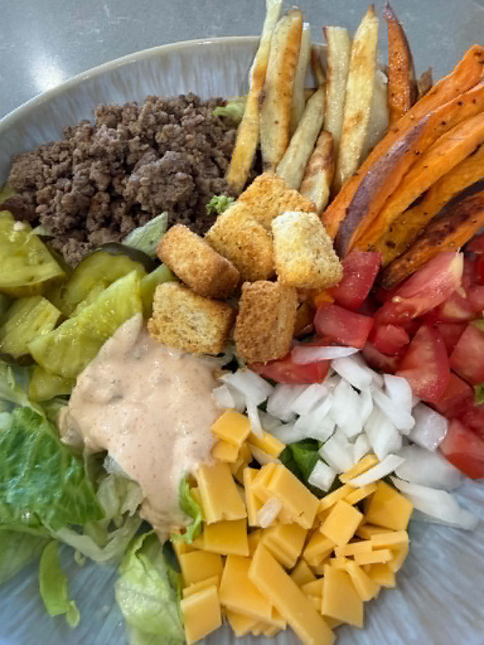 A bowl with ground beef, french fries, sweet potato fries, chopped tomatoes, chopped white onion, small slices of cheddar cheese, smash burger sauce, croutons, chopped lettuce, and pickle chips. 