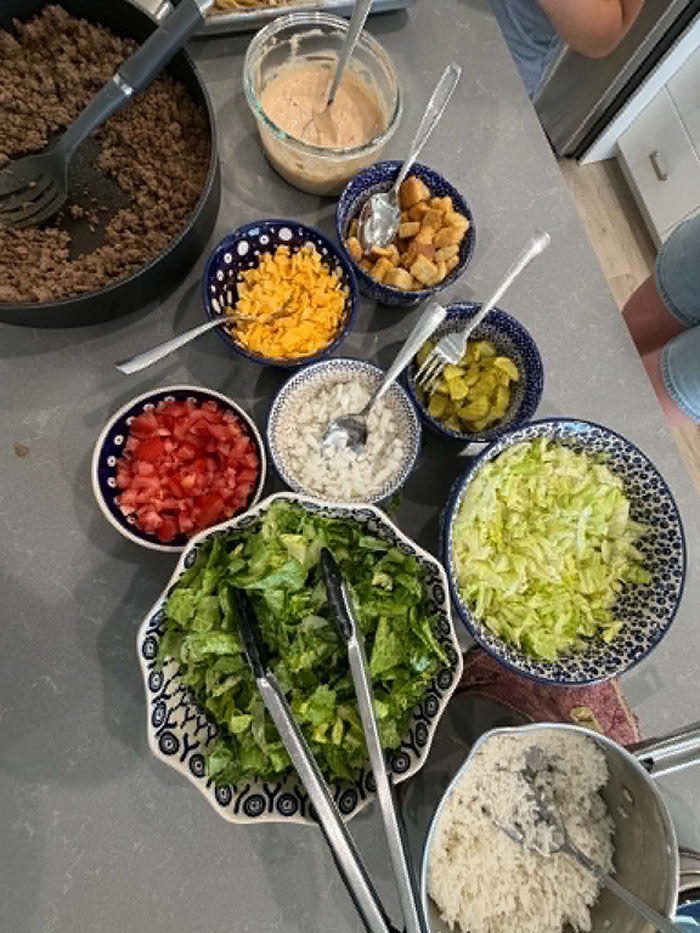 A pot of white rice with a serving spoon in it, a medium bowl with chopped romain lettuce and serving tongs, a medium bowl of chopped iceberg lettuce, a small bowl of diced tomatoes, a small bowl of diced onions with a spoon in it, a small bowl of sliced pickles with a fork in it, a small bowl of croutons with a spoon in it, a small bowl of small slices of cheddar cheese with a spoon in it, a medium measuring cup of burger sauce, and a large frying pan of ground beef with a serving spoon in it.