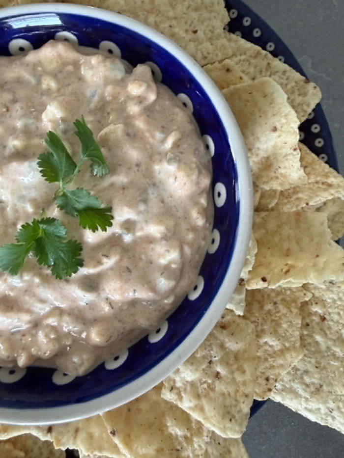 A partial picture of a small bowl of protein jalapeno dip with cilantro leaves on top in the middled sitting on a large plate surrounded by corn chips.