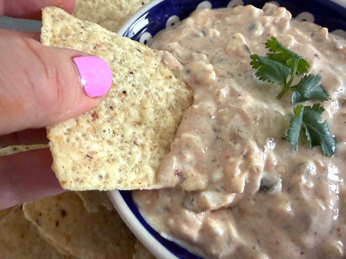 A small bowl of jalapeno dip with a few cilantro leaves on the top in the middle. A tortilla chip is partially inserted into the dip from the left.