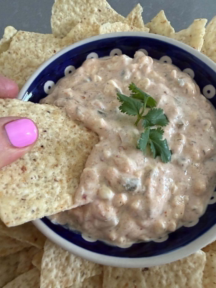 Jalapeno protein dip in a small bowl with a cilantro sprig on top in the middle, surrounded by tortilla chips. A tortilla chip held from the left is partly inserted into the dip.