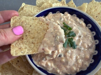 High protein jalapeno dip with a sprig of cilantro on top in the middle in a small bowl surrounded by corn chips. A corn chip with dip on the end is held above the bowl from the left.