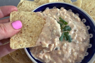 High protein jalapeno dip with a sprig of cilantro on top in the middle in a small bowl surrounded by corn chips. A corn chip with dip on the end is held above the bowl from the left.