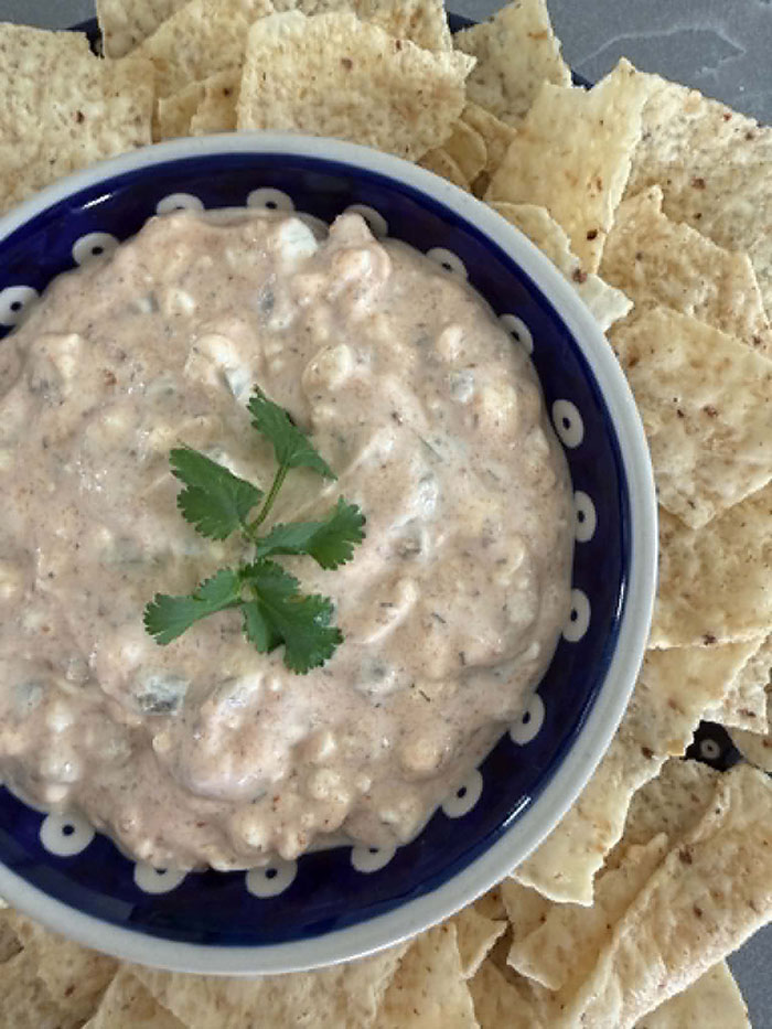 A small bowl filled with protein jalapeno dip with a sprig of cilantro on the top in the middle surrounded by tortilla chips.