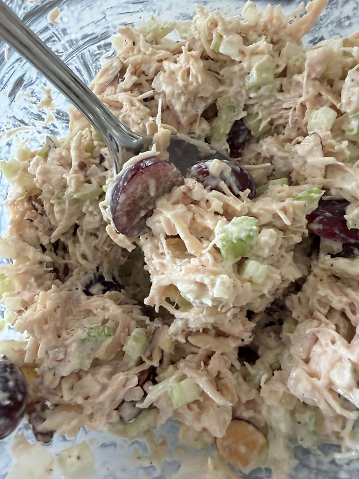 A glass bowl of chicken salad with grapes, celery, and red onion with a spoon inserted diagonally from the upper left corner.