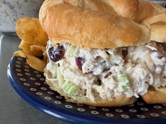 A croissant sandwich filled with extra protein chicken salad with barbecue chips sitting on a dark blue plate with a white polka-dotted pattern.