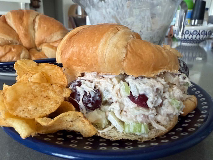 Barbecue chips and a croissant sandwich filled with extra protein chicken salad on a dark blue plate with white polka dots. Behind the plate is another plate with a croissant on it and a glass bowl of chicken salad.