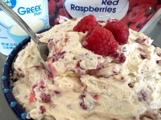 Frozen berry salad in a large bowl topped with three raspberries in the middle and a spoon inserted diagonally from the left. Behind the bowl is a carton of plain Greek yogurt and a bag of frozen red raspberries.