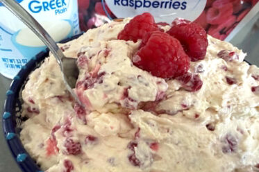 Frozen berry salad in a large bowl topped with three raspberries in the middle and a spoon inserted diagonally from the left. Behind the bowl is a carton of plain Greek yogurt and a bag of frozen red raspberries.