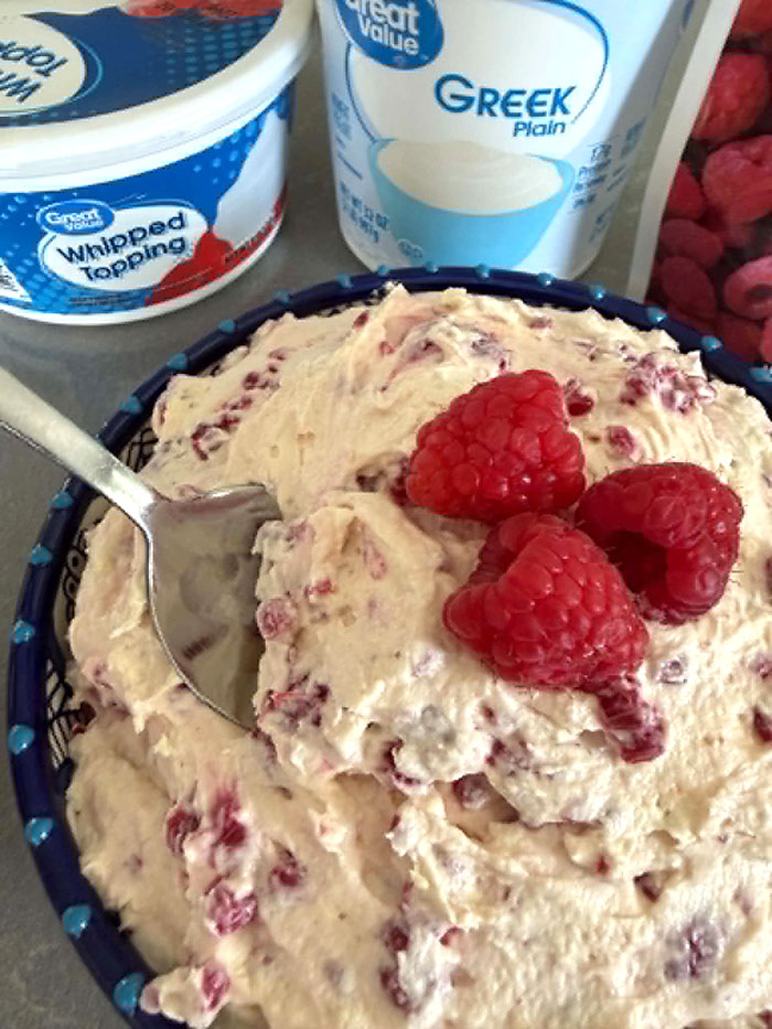 A bowl of Frozen Berry Salad with three whole raspberries on top in the middle and a spoon inserted diagonally from the left. Above the bowl are a whipped topping container and a plain Greek yogurt container.