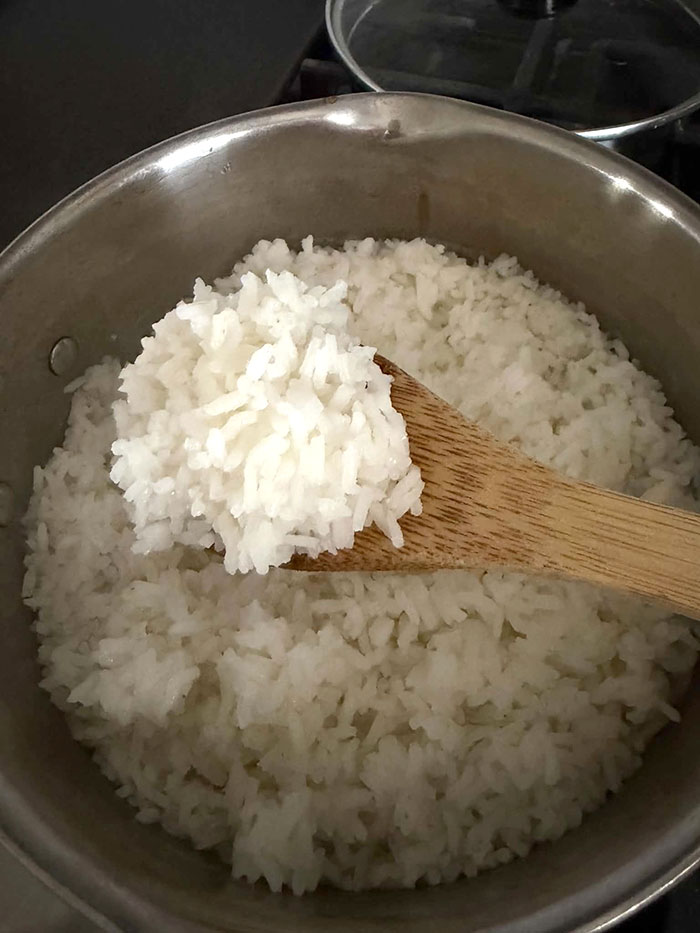 A pot of white, long-grain cooked rice with a wooden spoon with rice on it held above from the right.