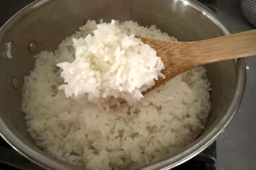 A wooden spoon with cooked long-grain rice on it held above a pot of cooked white long-grain rice from the right.