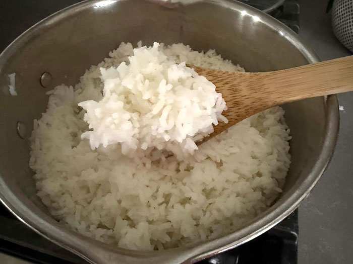 A wooden spoon with cooked long-grain rice on it held above a pot of cooked white long-grain rice from the right. 