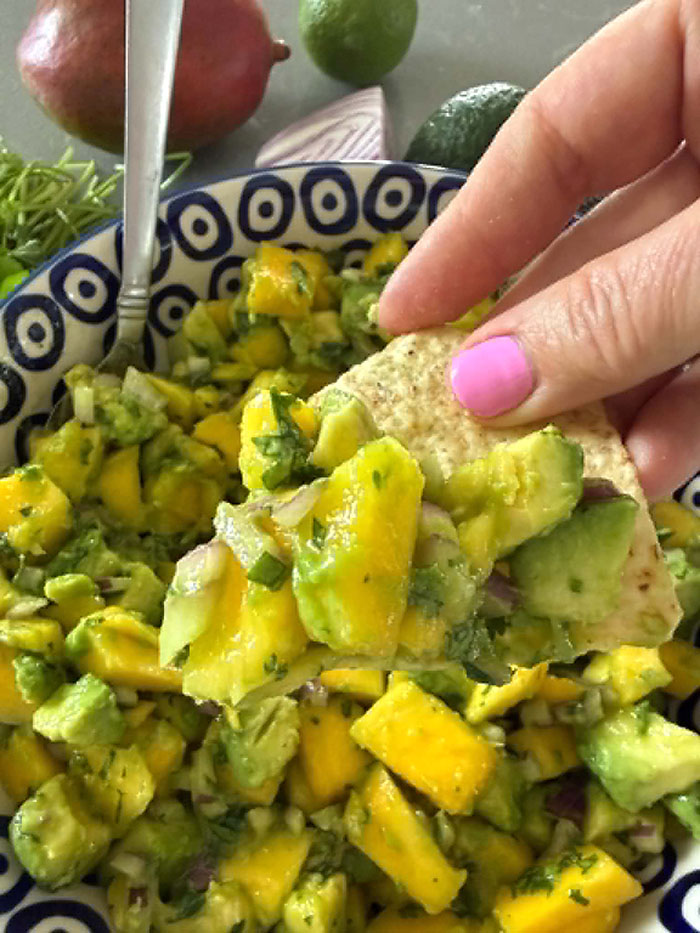 Mango Avocado Salsa in a bowl with a white background and blue circles with a dot in the middle. A tortilla chip with salsa is held above the bowl. A spoon is standing upright in the salsa on the upper left. At the top is a mango, two limes, half a red onion, and some cilantro.