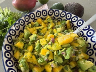 A white bowl with blue circles and a dot in the middle filled with Mango Avocado Salsa. A spoon is inserted in the salsa from the right. At the top is an avocado, lime, red onion slice, a mango, and a cilantro bunch.