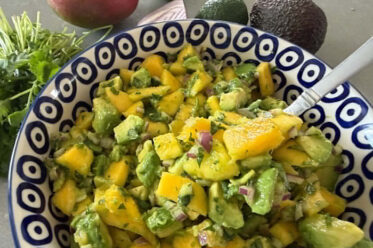 A white bowl with blue circles and a dot in the middle filled with Mango Avocado Salsa. A spoon is inserted in the salsa from the right. At the top is an avocado, lime, red onion slice, a mango, and a cilantro bunch.