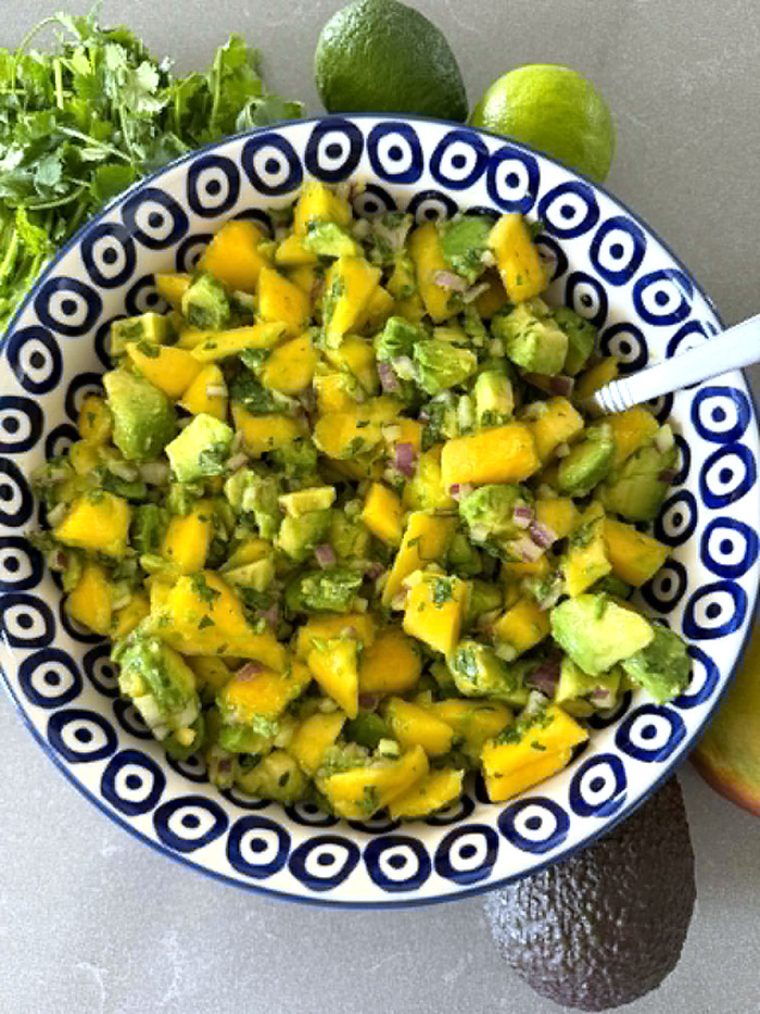 Mango Avocado Salsa in a white bowl with blue circles with a dot in the middle. A spoon is inserted into the salas from the right. In the bottom right corner is an avocado. At the top is a cilantro bunch and two limes.