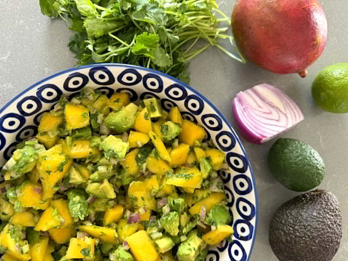 A white bowl with blue circles with a dot in the middle of each circle filled with Mango Avocado Salsa. To the right of the both is a half a red onion, two limes, and an avocado. In the upper right corner is a mango. To the left of the mango is a cilantro bunch.