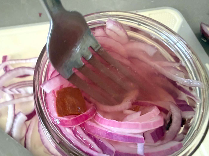 An opened jar of red pickled onions with a fork inserted diagonally from the top middle. In the background are sliced red onions on a cutting board.